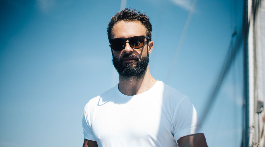 Bearded man facing viewer standing on white yacht wearing sunglasses and white tshirt on bright sunny day