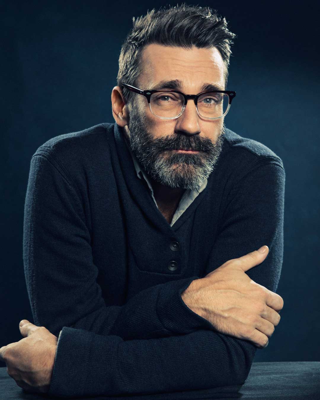 Bearded man wearing navy cardigan and gradient two tone glasses frame in dark room