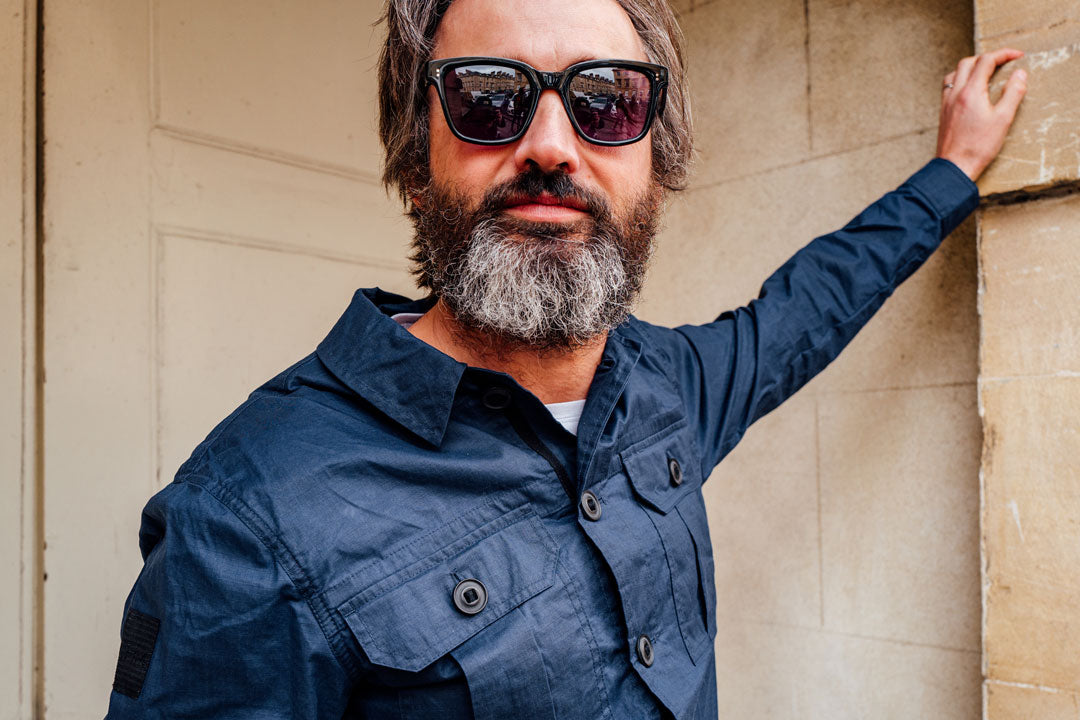 Bearded man with long hair wearing blue jacket and square black sunglasses frame