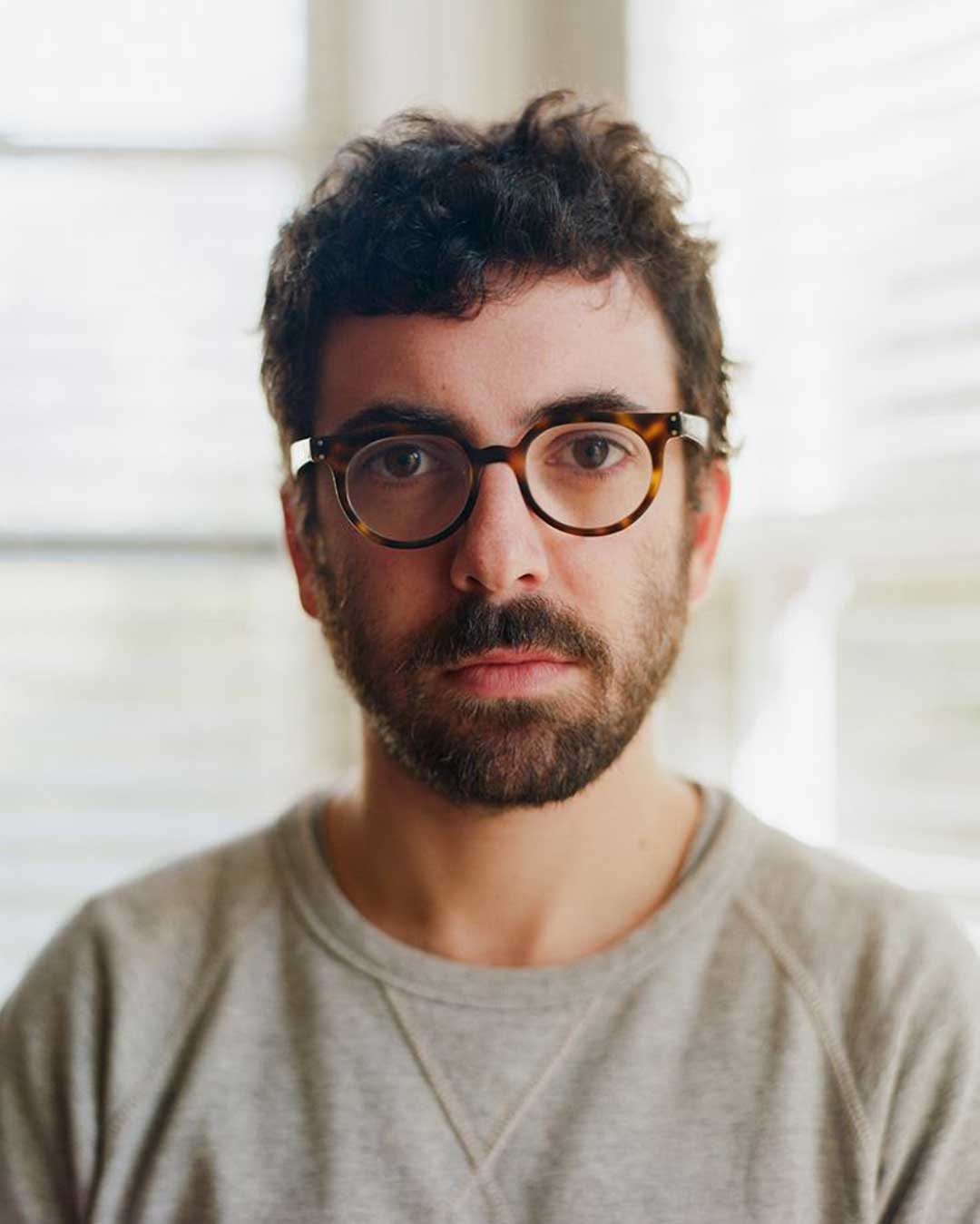 Bearded man with short curly hair wearing rounded square tortoise glasses and grey sweater