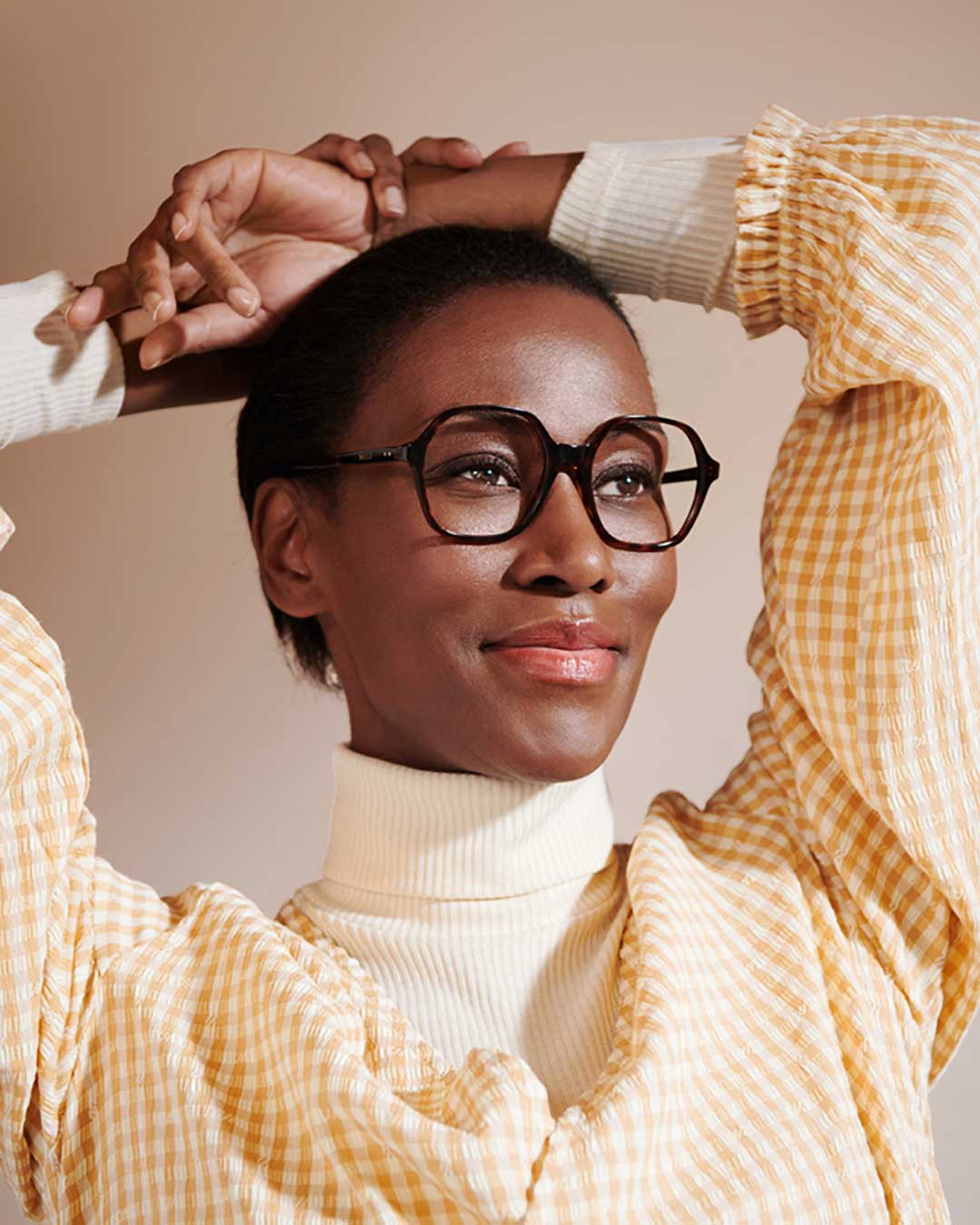 Black woman with short hair with her hands on her head smiling wearing oversized eyeglasses