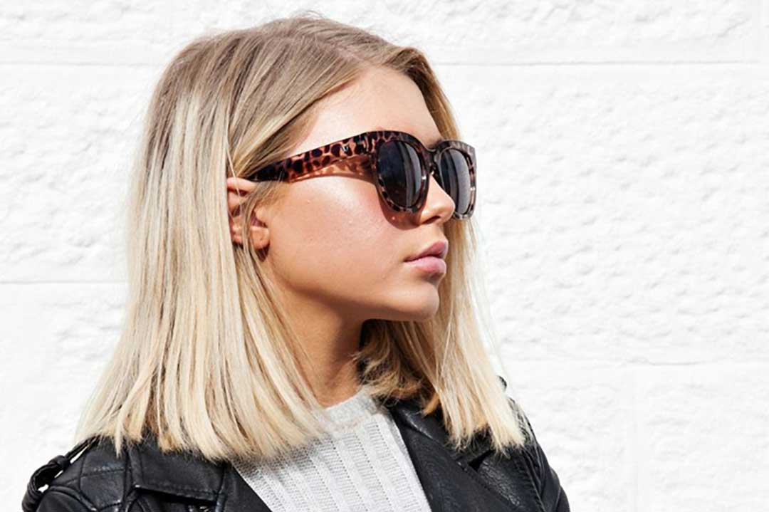 Blonde female wearing large tortoise shell sunglasses standing in front of white wall