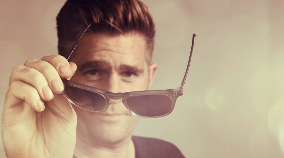 Blonde haired man presenting a pair of grey sunglasses to the viewer