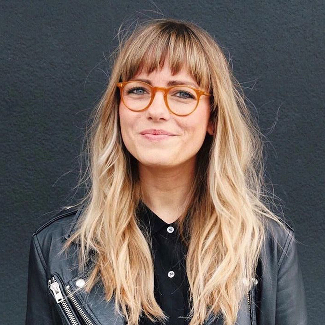 Blonde lady with bangs wearing round amber glasses and black leather jacket smiling