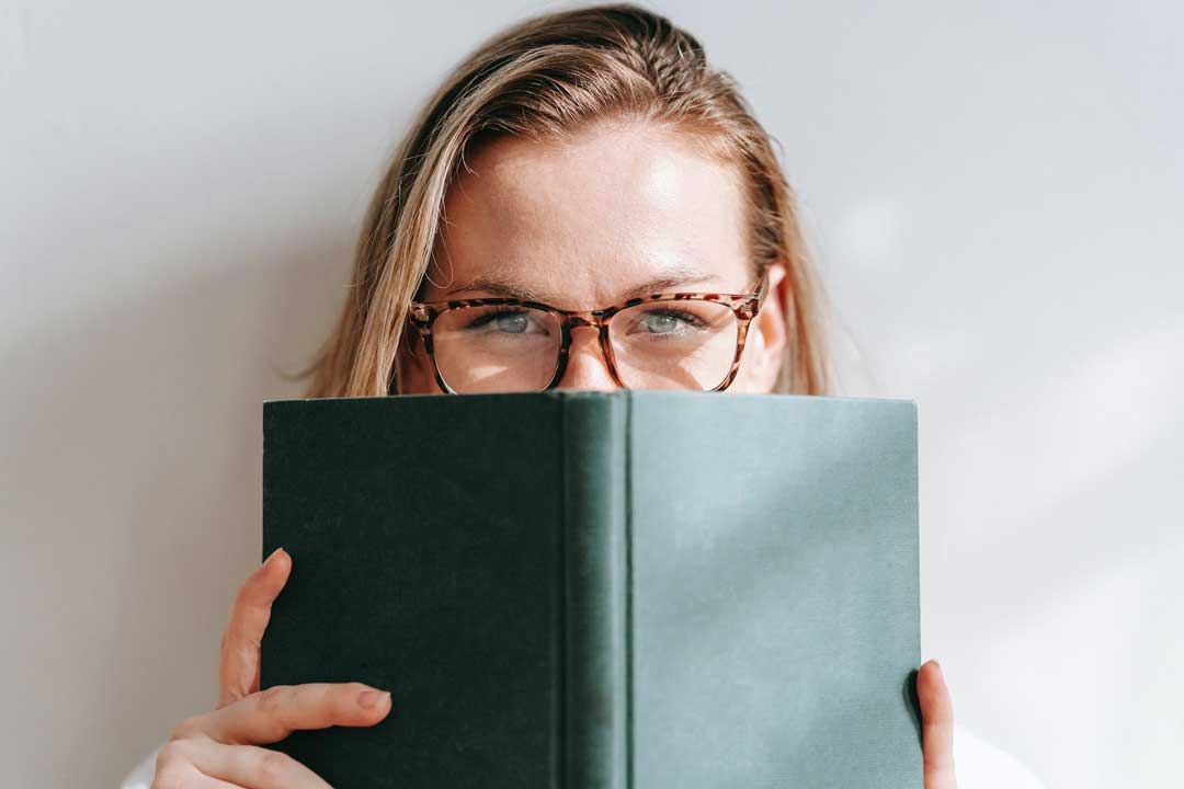 Blonde woman wearing tortoise shell eyeglasses covering her mouth with a green book