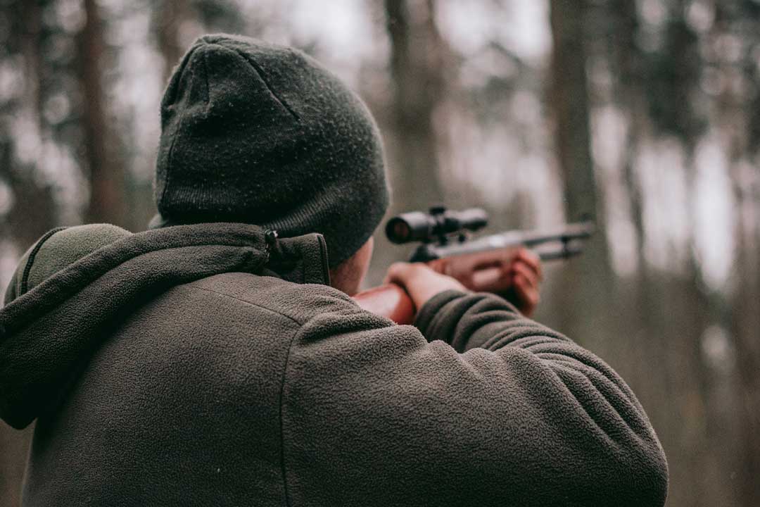 Close view of man firing a rifle in woodland wearing hat and thick jumper