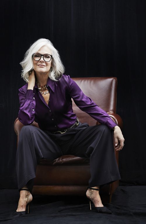 Confident mature lady sitting in a purple chair with purple blouse and prominent dark spectacles smiling at camera
