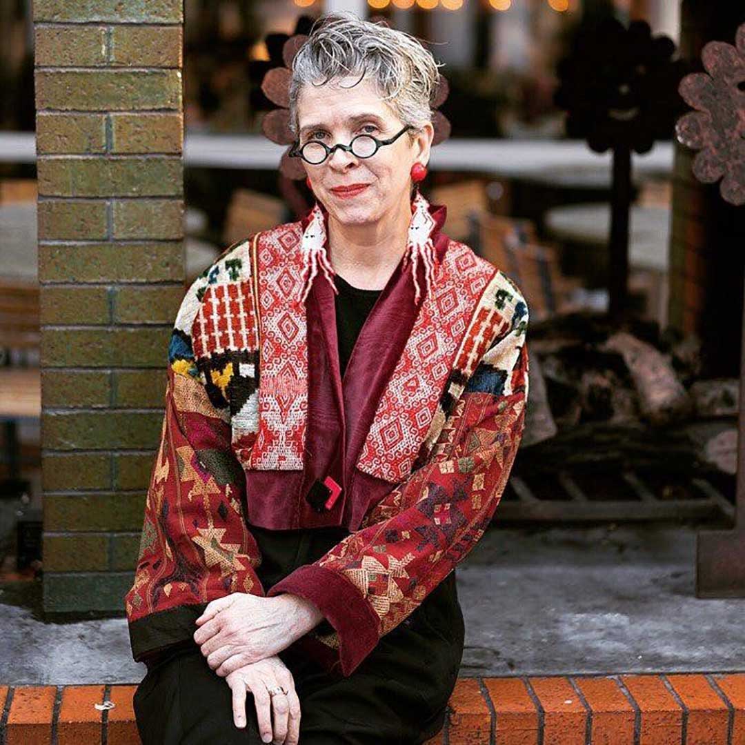 Eccentric looking woman sitting on a wall with curly pixie haircut and undersized round spectacles