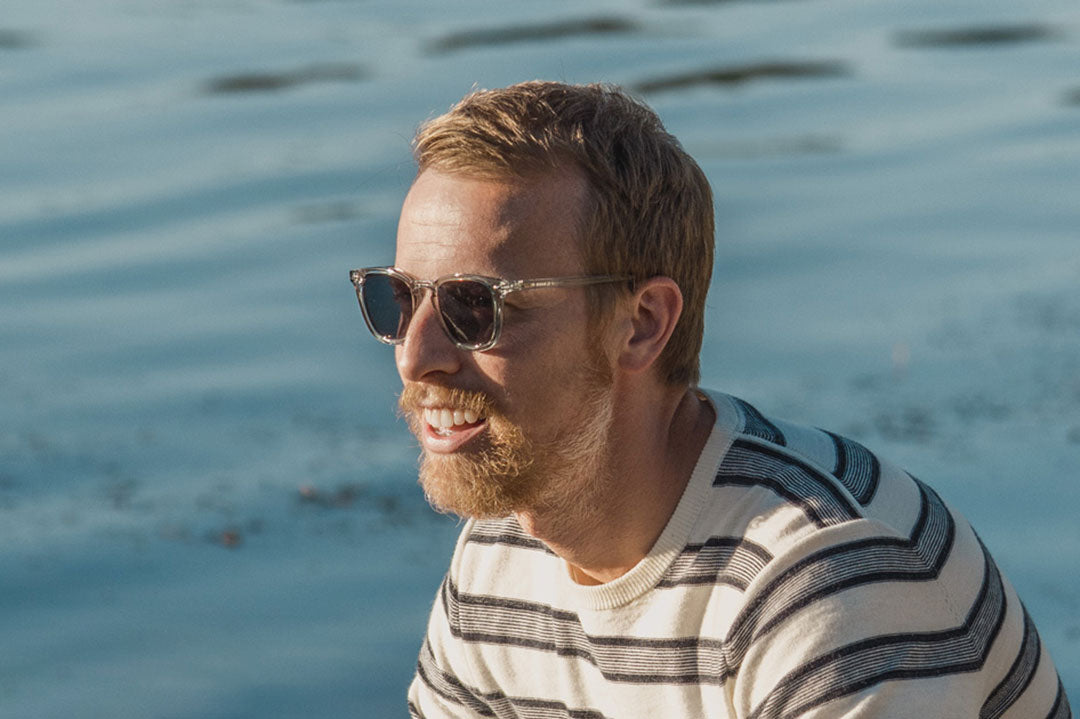 Ginger haired man wearing clear frame sunglasses beside water on sunny day