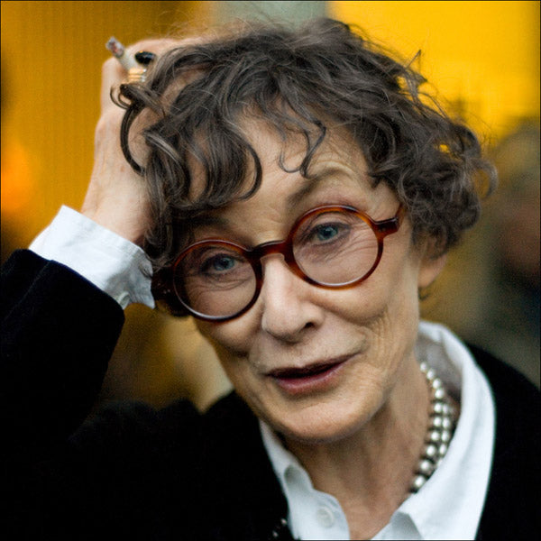 Grey haired lady with round amber glasses frame with one hand on her head holding a cigarette