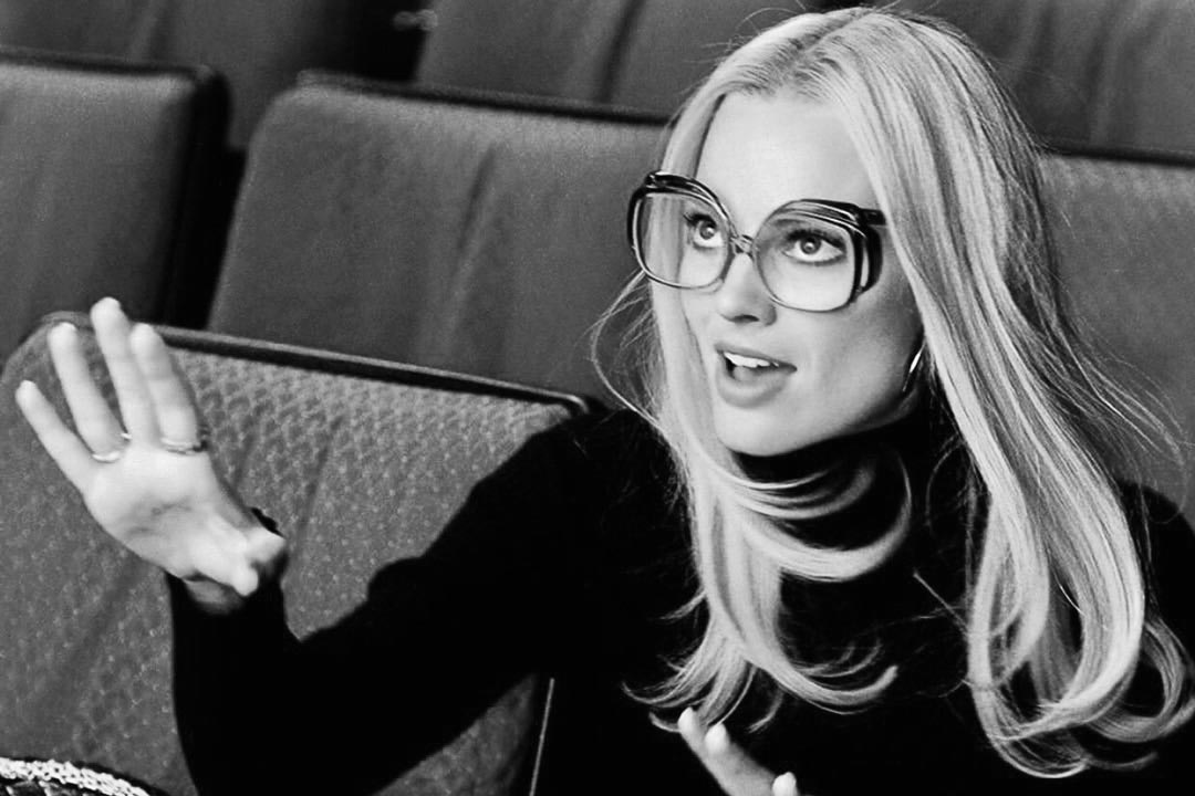 Grey scale image of actress Margot Robbie sitting in cinema seats wearing large oversized eyeglasses
