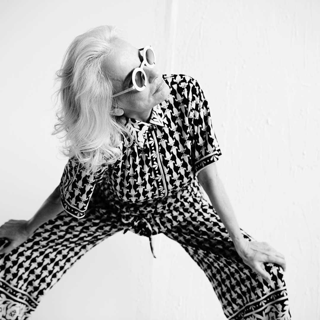 Lady wearing dogtooth suit and trousers with round white sunglasses frame and long grey hair