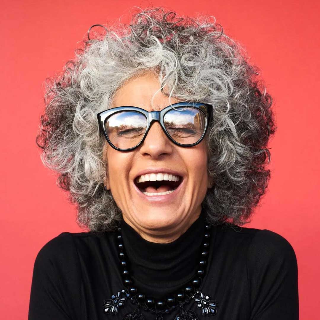 Lady with big curly afro haircut laughing wearing thick black cat eye glasses frame in front of pink background