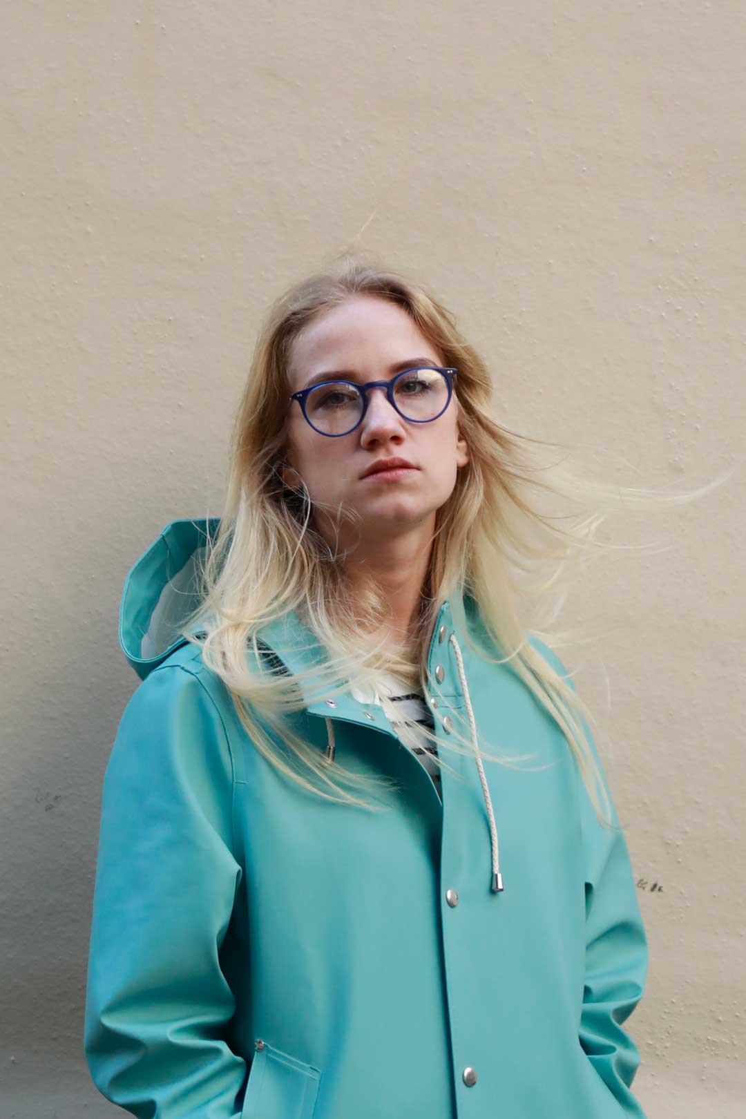 Lady with light blonde hair standing in front of wall wearing light blue jacket and round blue eyeglasses frame