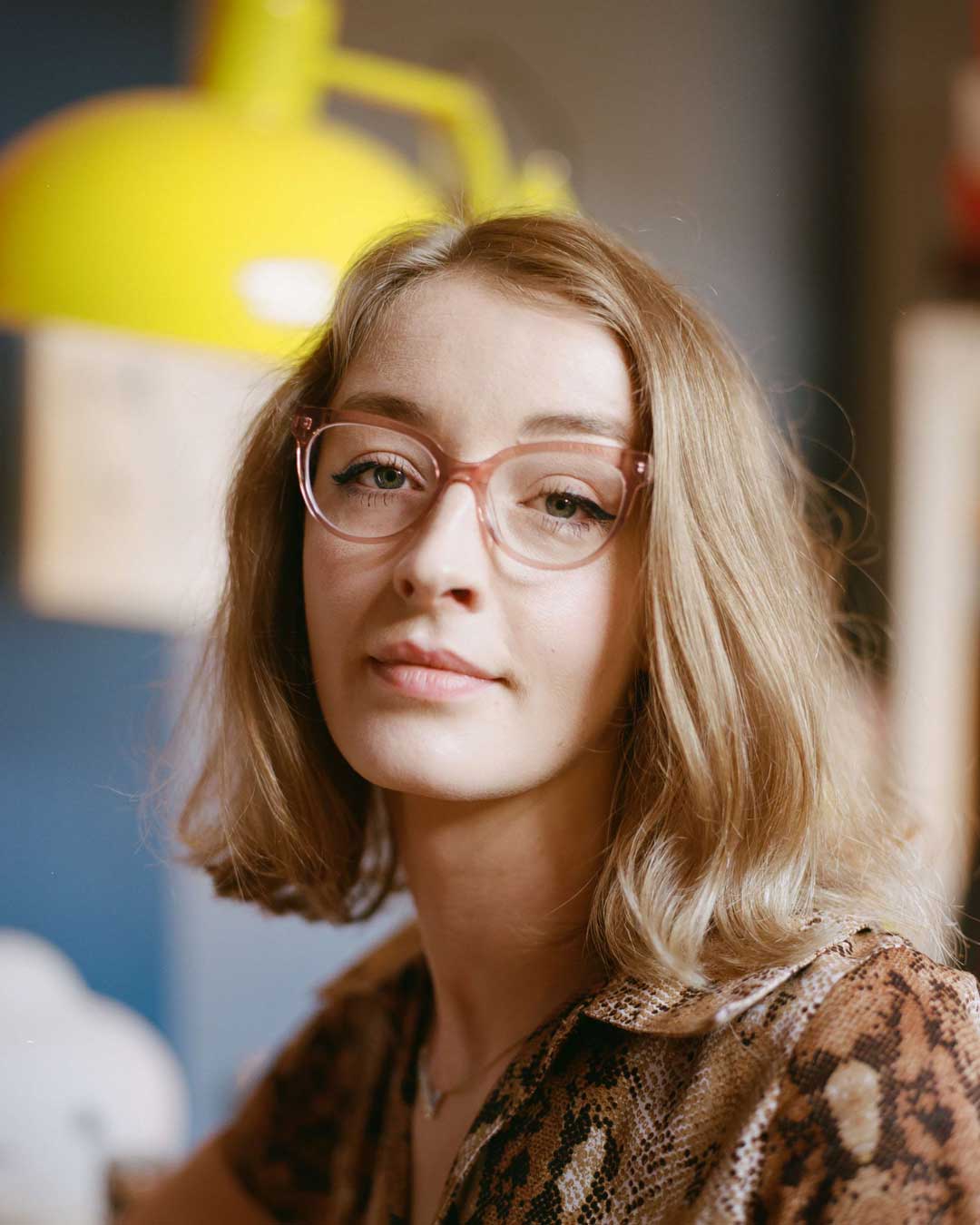 Lady with long blonde hair sitting beside yellow lamp wearing pink crystal cat eye glasses frame