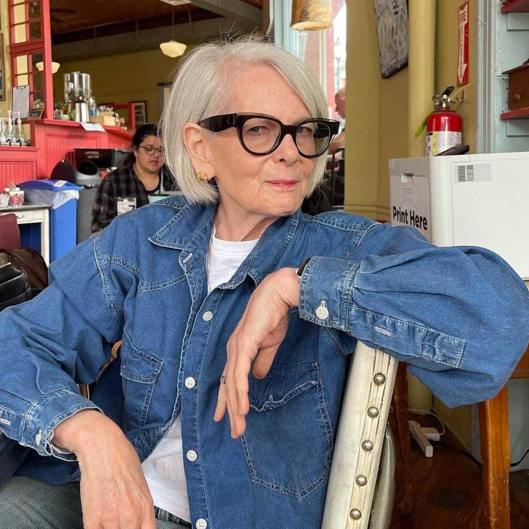 Lady with short grey haircut sitting in café wearing blue denim jacket and very thick black glasses