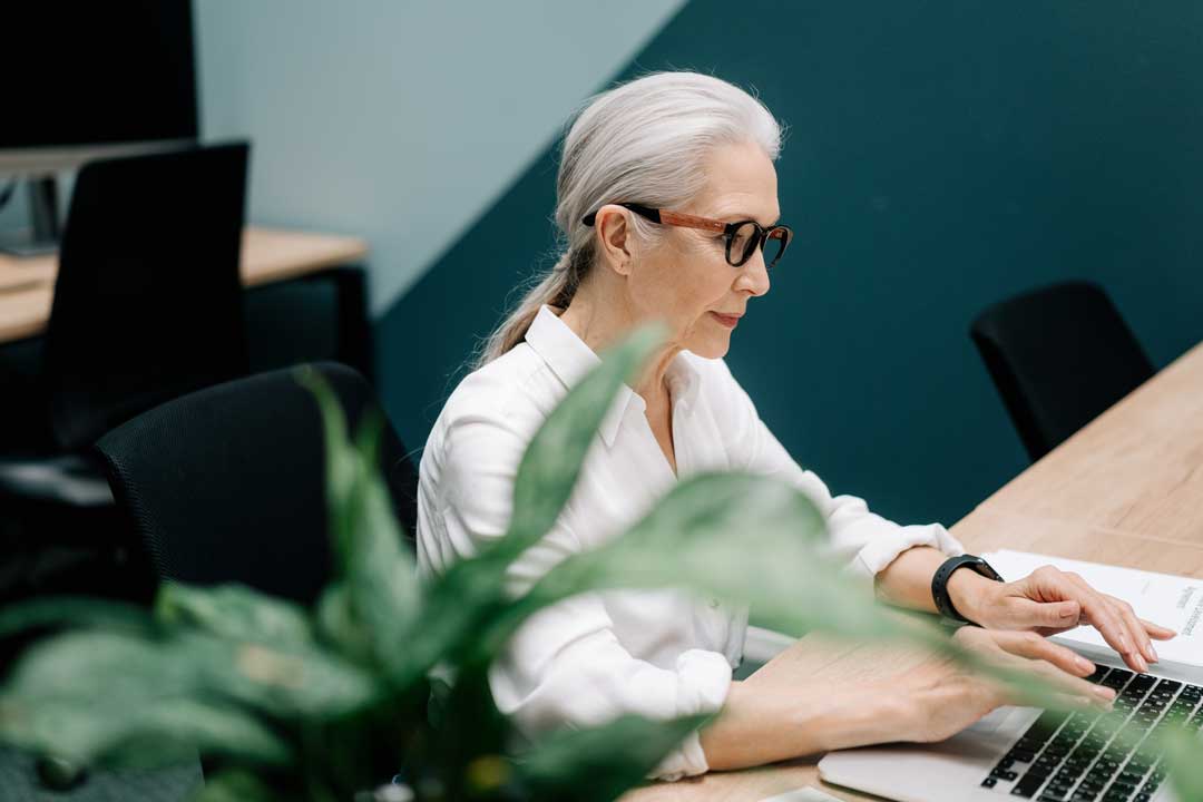 Lady with tied back long grey hair sitting at computer wearing white shirt and chunky eyeglasses