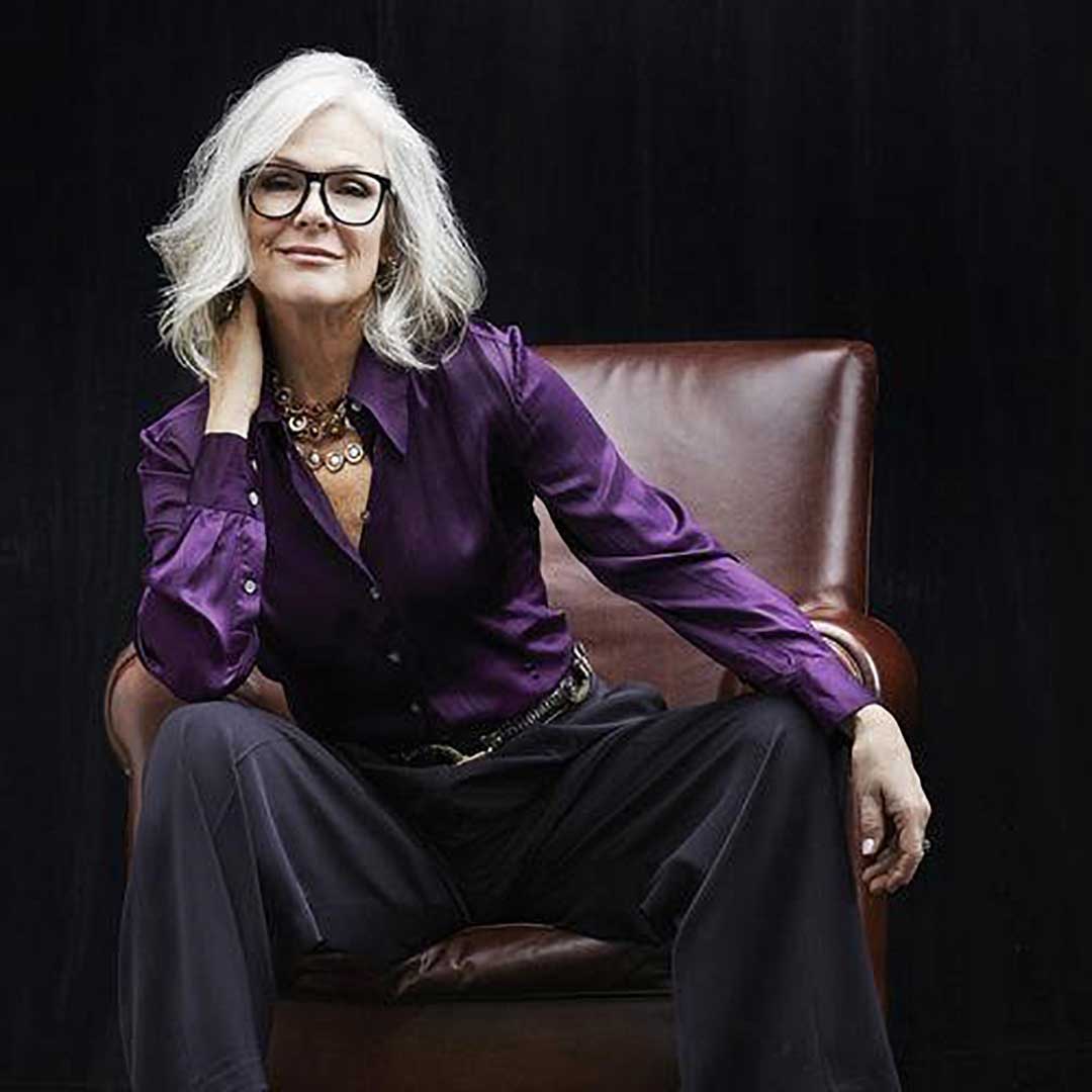 Lady with wavy bob haricut sitting in brown leather chair wearing purple top and square black eyeglasses