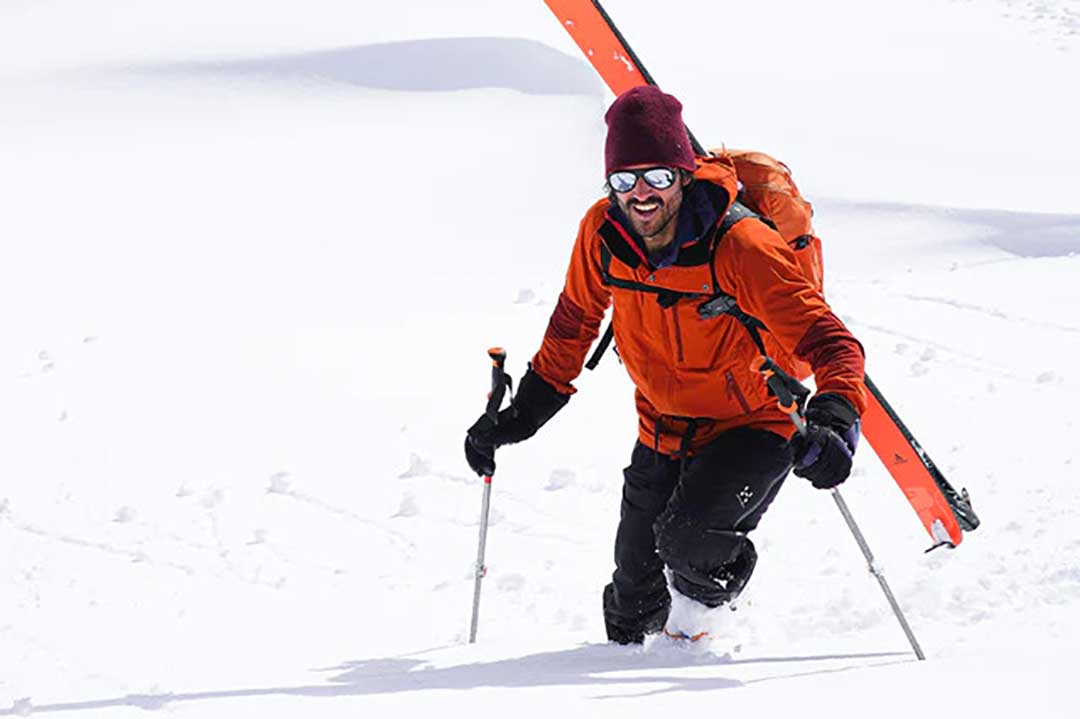 Man hiking up snow hill with skis on his back wearing mirrored skiing sunglasses