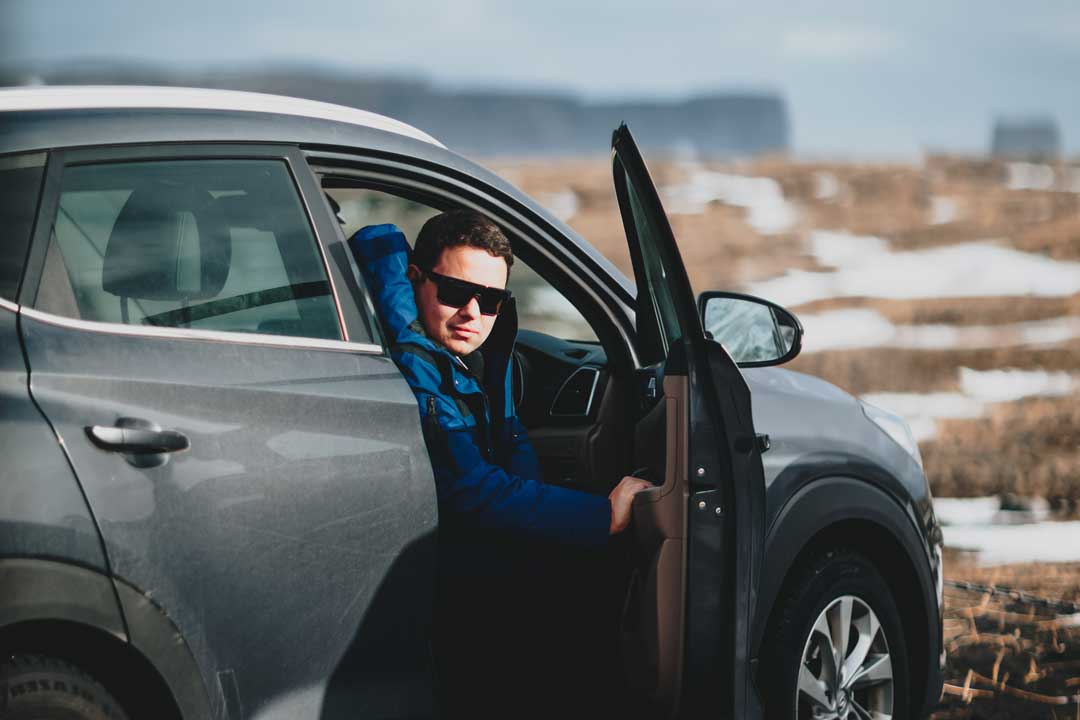 Man in black estate car wearing blue jacket and square black sunglasses frame on cold winter day