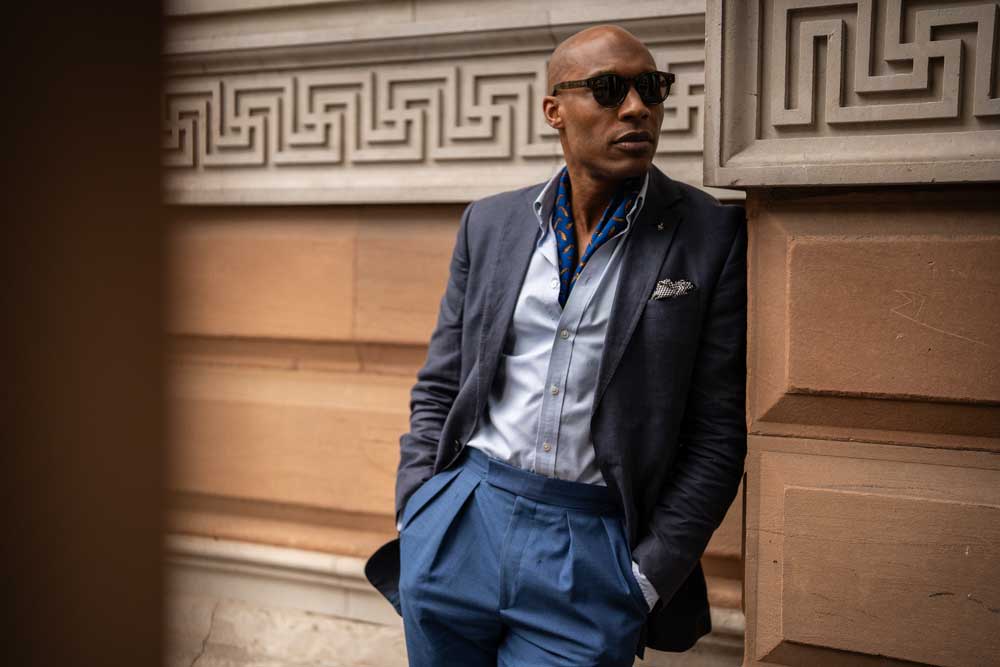 Man wearing blue suit and sunglasses leaning against wall in street