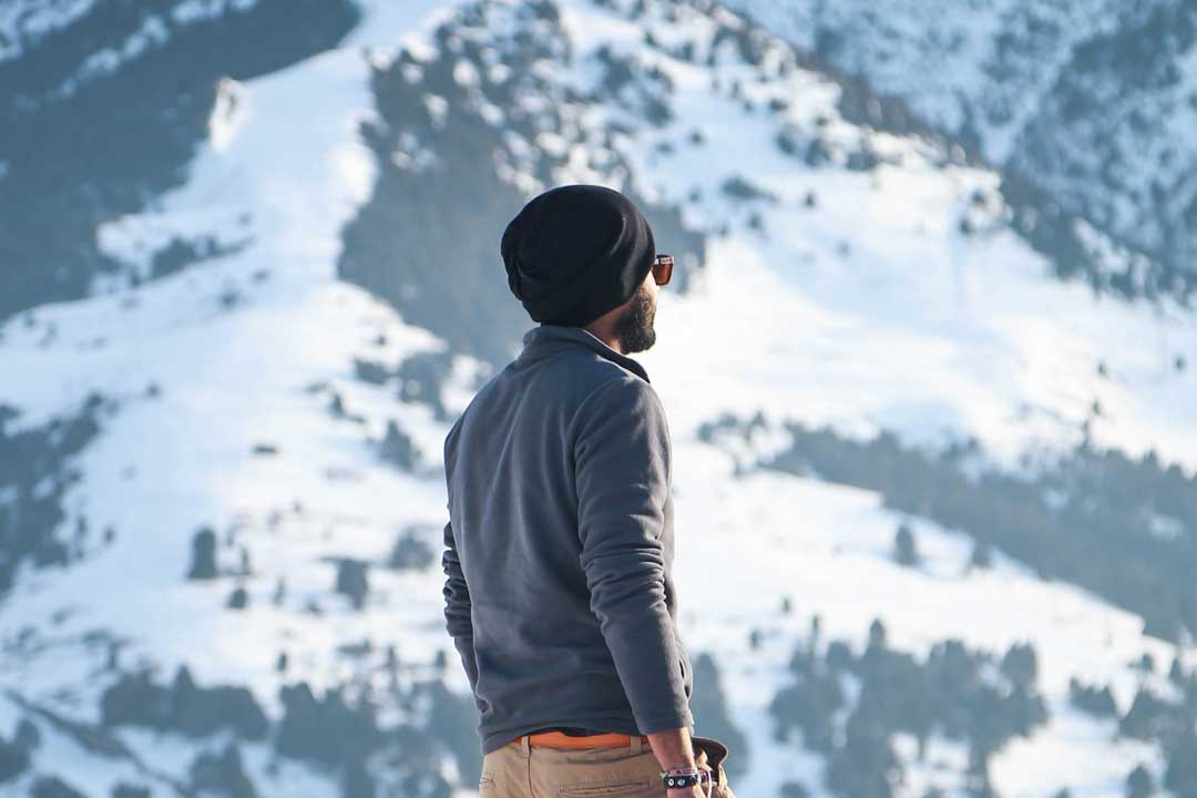 Man wearing hat and sunglasses frame looking towards snow covered mountains