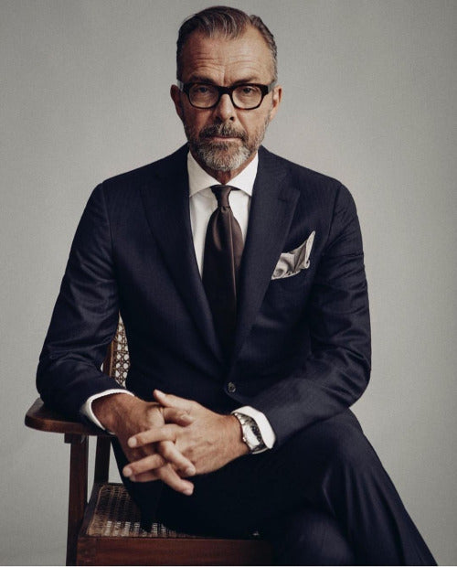 Man wearing suit and tie thick dark glasses frame sitting in a chair with his hands on the arm