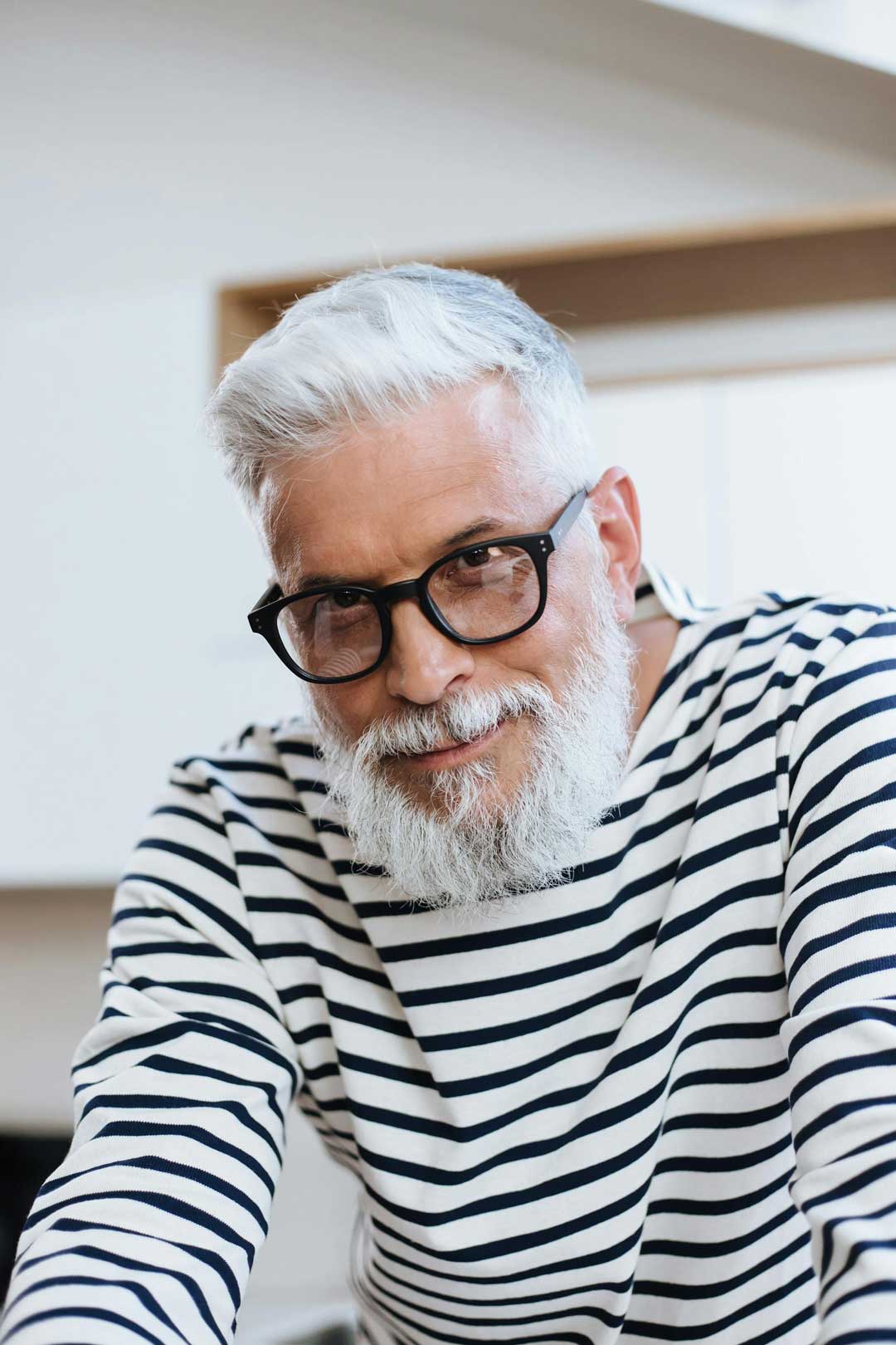 Mature beareded man with grey hair wearing thick oval eyeglasses and striped sweater