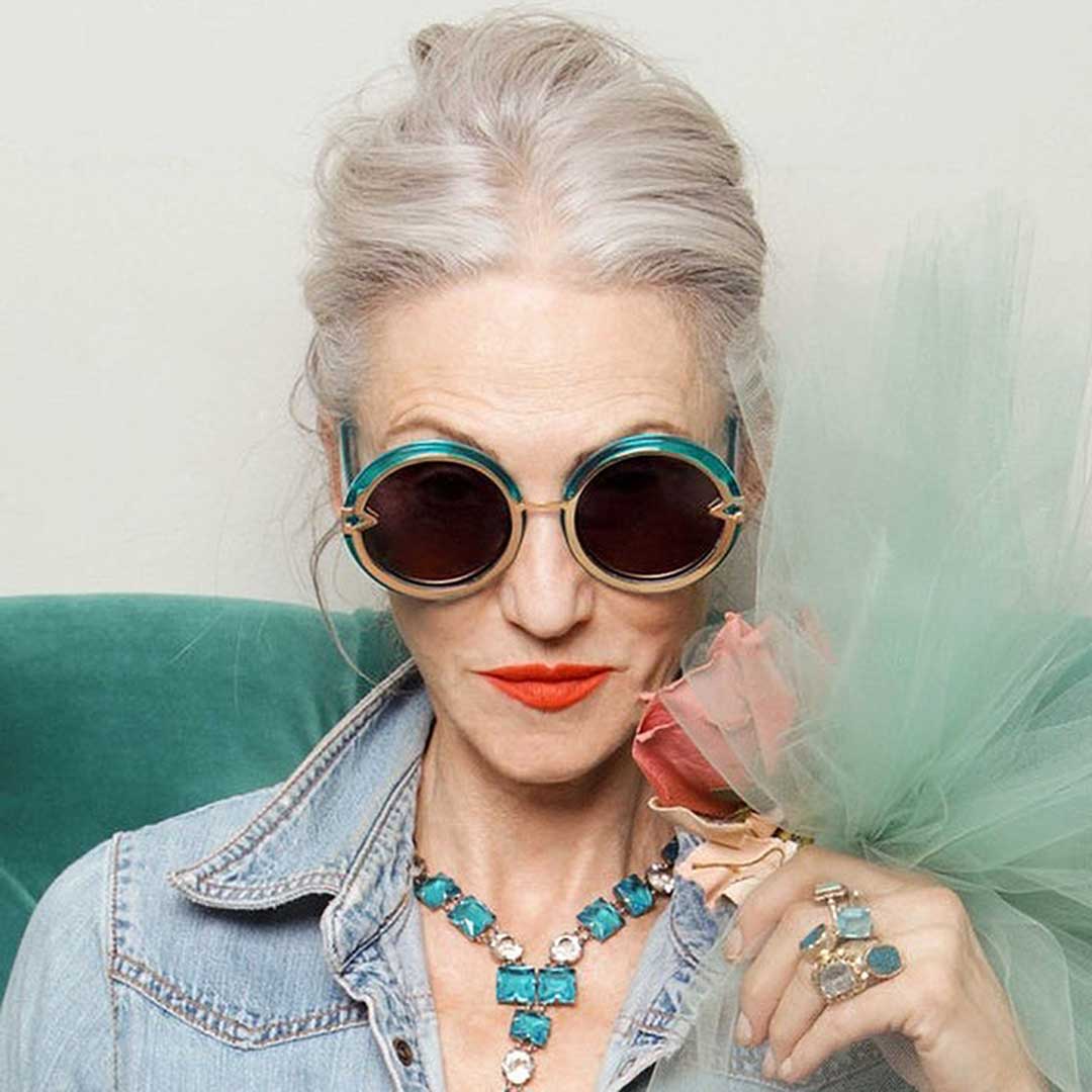 Mature female with long grey hair tied up wearing round teal and gold sunglasses frame