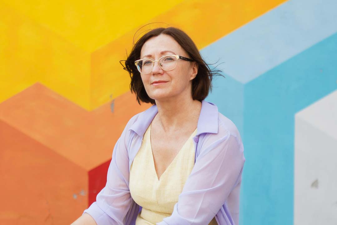 Mature lady wearing cat eye glasses frame sitting in front of colourful wall