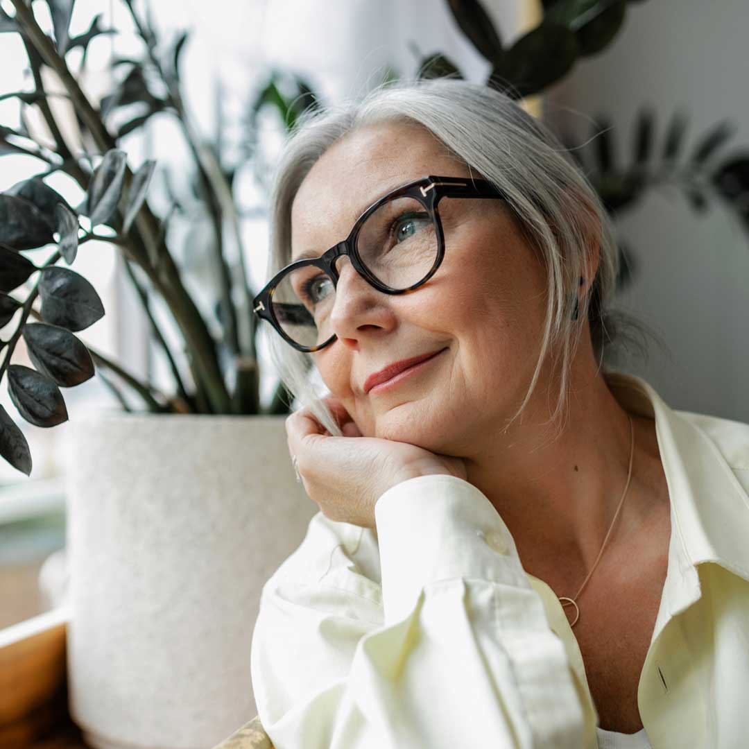 Mature lady with long grey hair and tortoise shell eyeglasses looking out of window