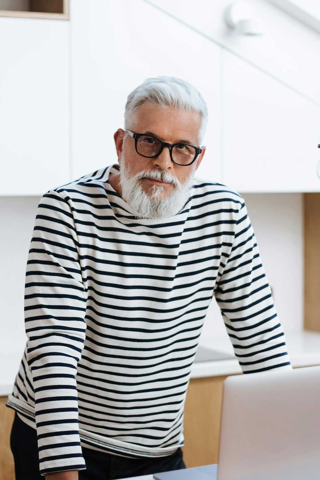 Mature man standing in kitchen beside laptop computer wearing thick square eyeglasses frame and striped jumper
