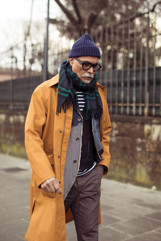 Mature man wearing bright yellow jacket blue hat glasses walking down the street looking away from the viewer