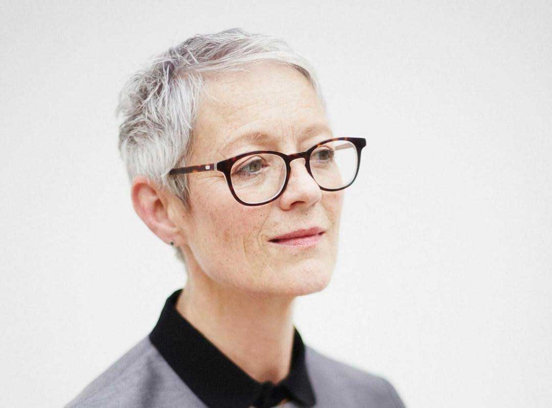 Mature woman with very short grey hair wearing rectangular eyeglasses in front of white background