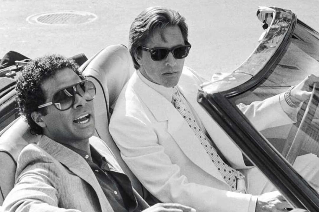 Miami Vice characters Rico and Sonny sitting in an open top sports car wearing suits and sunglasses on sunny day