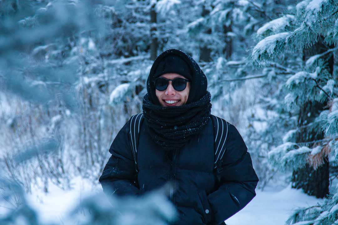 Person standing in forest on cold winter day wearing hat coat and round black sunglasses frame