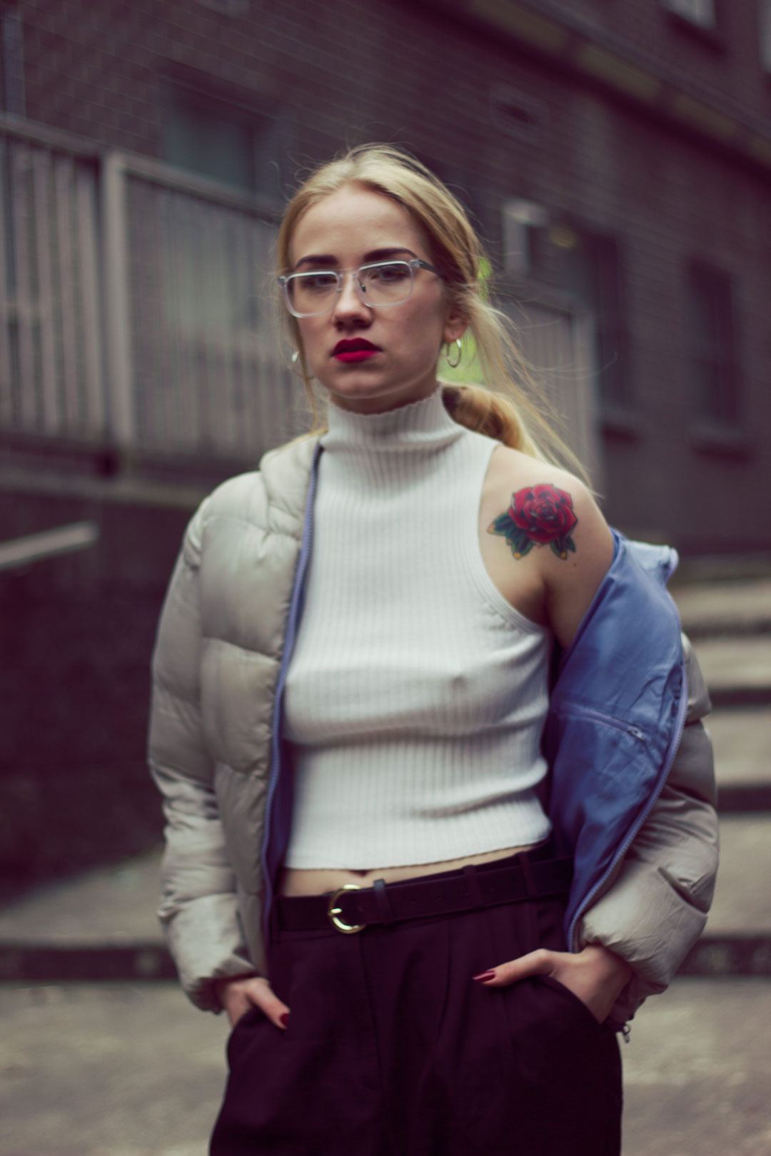 Portrait of young blonde woman in street wearing puff jacket and grey eyeglasses staring directly at viewer