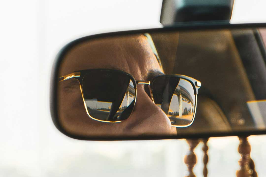 Reflection of person wearing Clubmaster sunglasses in car rear view mirror