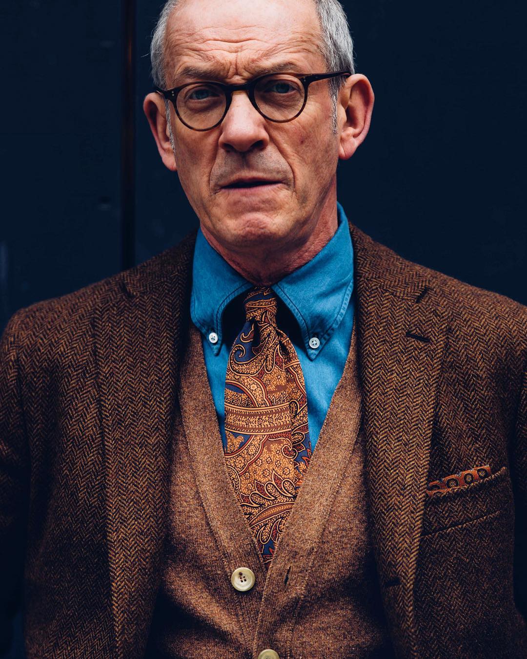 Serious looking man with blue shirt and brown suit jacket wearing round tortoise spectacle frames