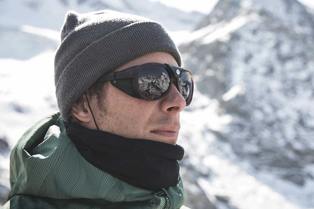 Side view of man standing in the snowy mountains wearing hat and side shield sunglasses frame