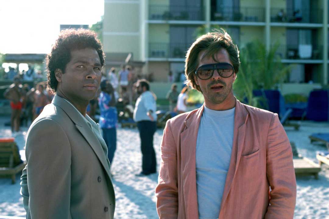 Sonny Crockett wearing pink suit and Aviator sunglasses on beach standing beside Rico Tubbs in Miami Vice episode 1