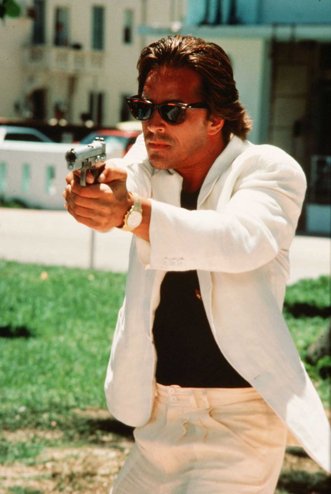 Sonny Crockett wearing white suit and RayBan Wayfarer sunglasses holding a gun ready to shoot