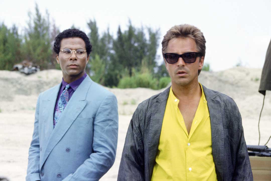 Sonny from Miami Vice wearing black Persol Ratti 69218 sunglasses frame and suit standing beside his partner Rico Tubbs