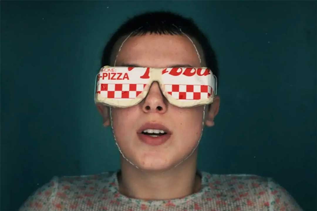 Stranger Things character Eleven floating in water wearing block out sunglasses made from a pizza box