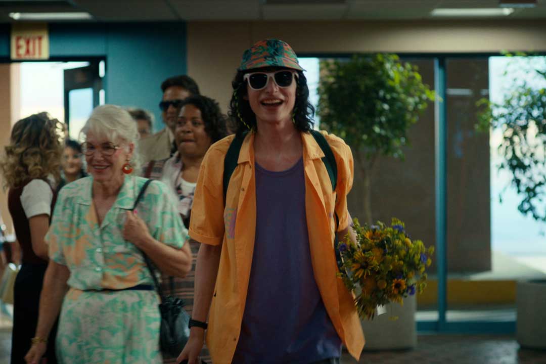 Stranger Things character Mike Wheeler walking through airport arrivals hall wearing colourful shirt and sunglasses smiling carrying flowers
