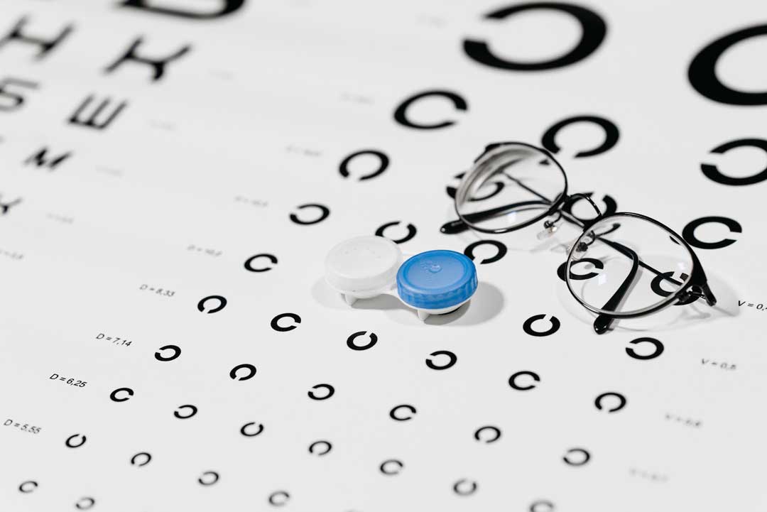 Thin black spectacles lying folded upon white eye test chart beside contact lenses