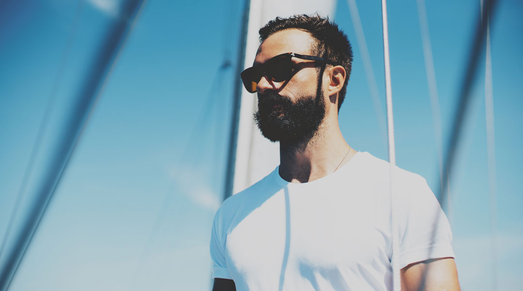 Three quarter view of bearded man on sail yacht wearing polarised sunglasses and white shirt