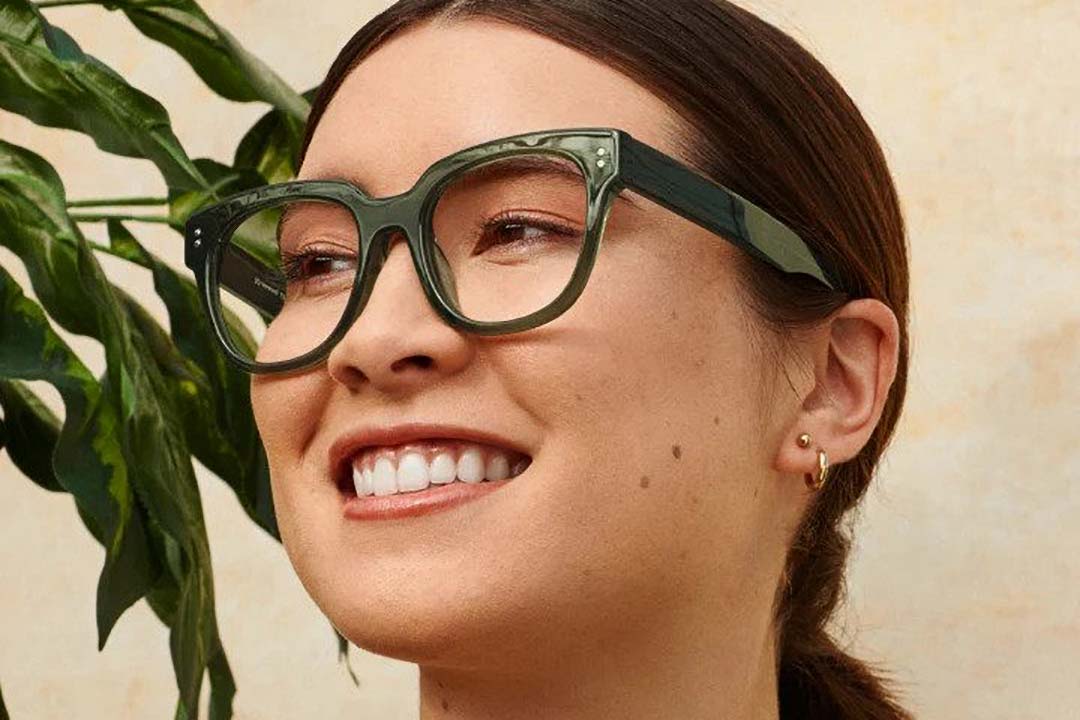 Three quarter view of lady wearing thick green eyeglasses smiling standing beside house plant