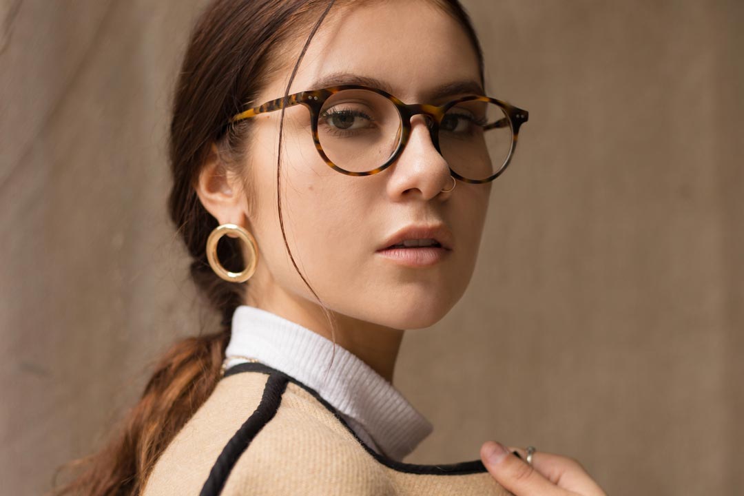 Three quarter view of young lady wearing round tortoise eyeglasses in front of brown wall