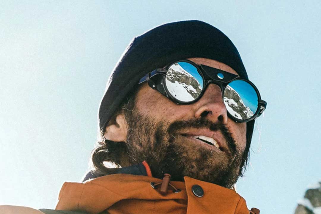 Upward view of man wearing round glacier sunglasses frame with side shields and orange jacket in mountains on a bright sunny day